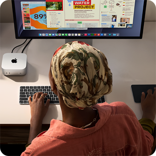 Íróasztalnál ülő ember egy Mac minin gépel, melyet Studio Displayjel, Magic Keyboarddal és Magic Trackpaddel használ, körülötte különféle tárgyak