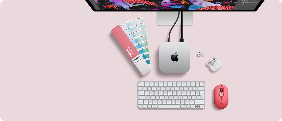Mac mini felülnézetből, ezüstszínű alumíniumházzal, lekerekített sarkokkal, középen fényes fekete Apple-logóval, kábellel csatlakoztatva, a képen felül egy Studio Display egy része, kinyitott fedelű AirPods 4, Pantone-színminták, számbillentyűzetes billentyűzet és egér