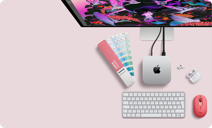 Mac mini felülnézetből, ezüstszínű alumíniumházzal, lekerekített sarkokkal, középen fényes fekete Apple-logóval, kábellel csatlakoztatva, a képen felül egy Studio Display egy része, kinyitott fedelű AirPods 4, Pantone-színminták, számbillentyűzetes billentyűzet és egér